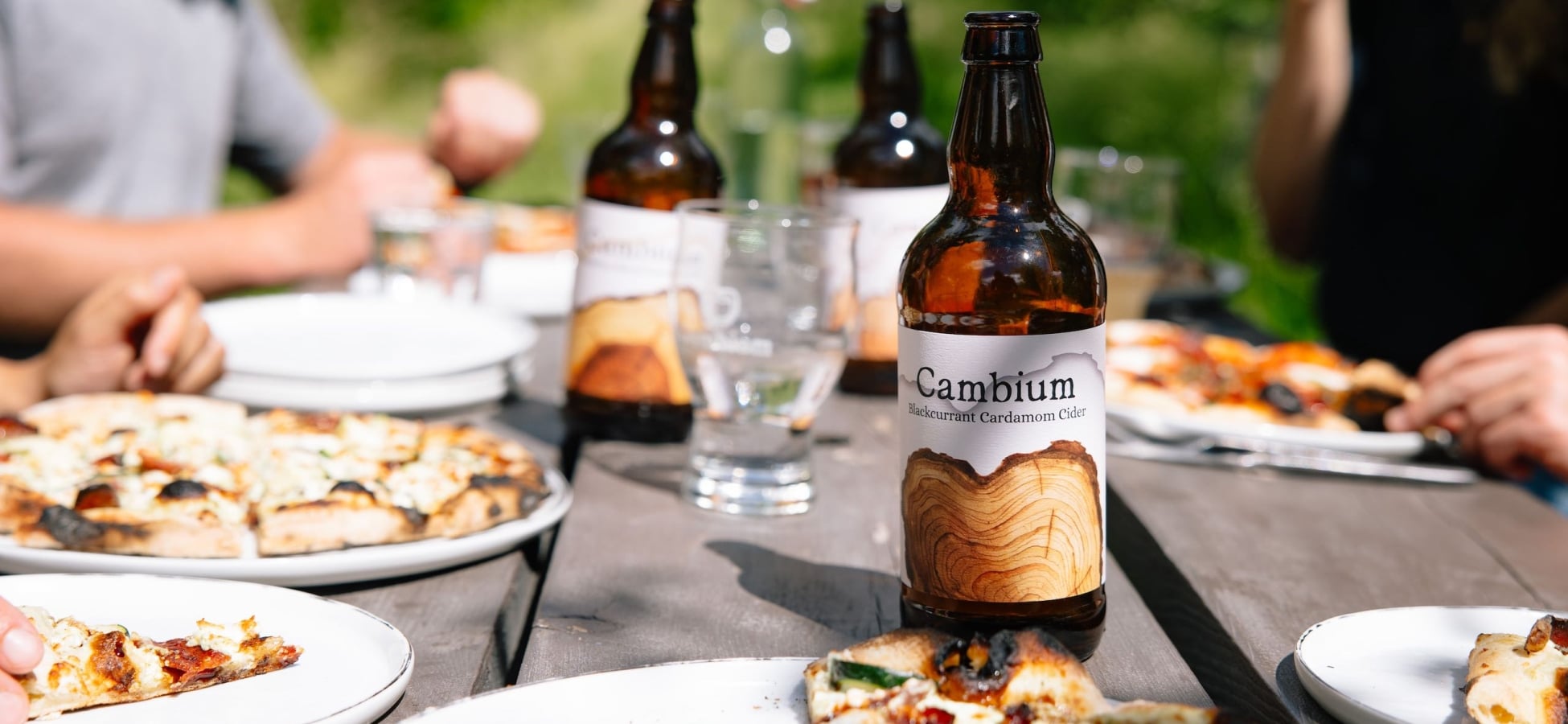 Close up of a Cambium Cider Co. cider bottle on a picnic table with pizza, people, and more cider in the background.