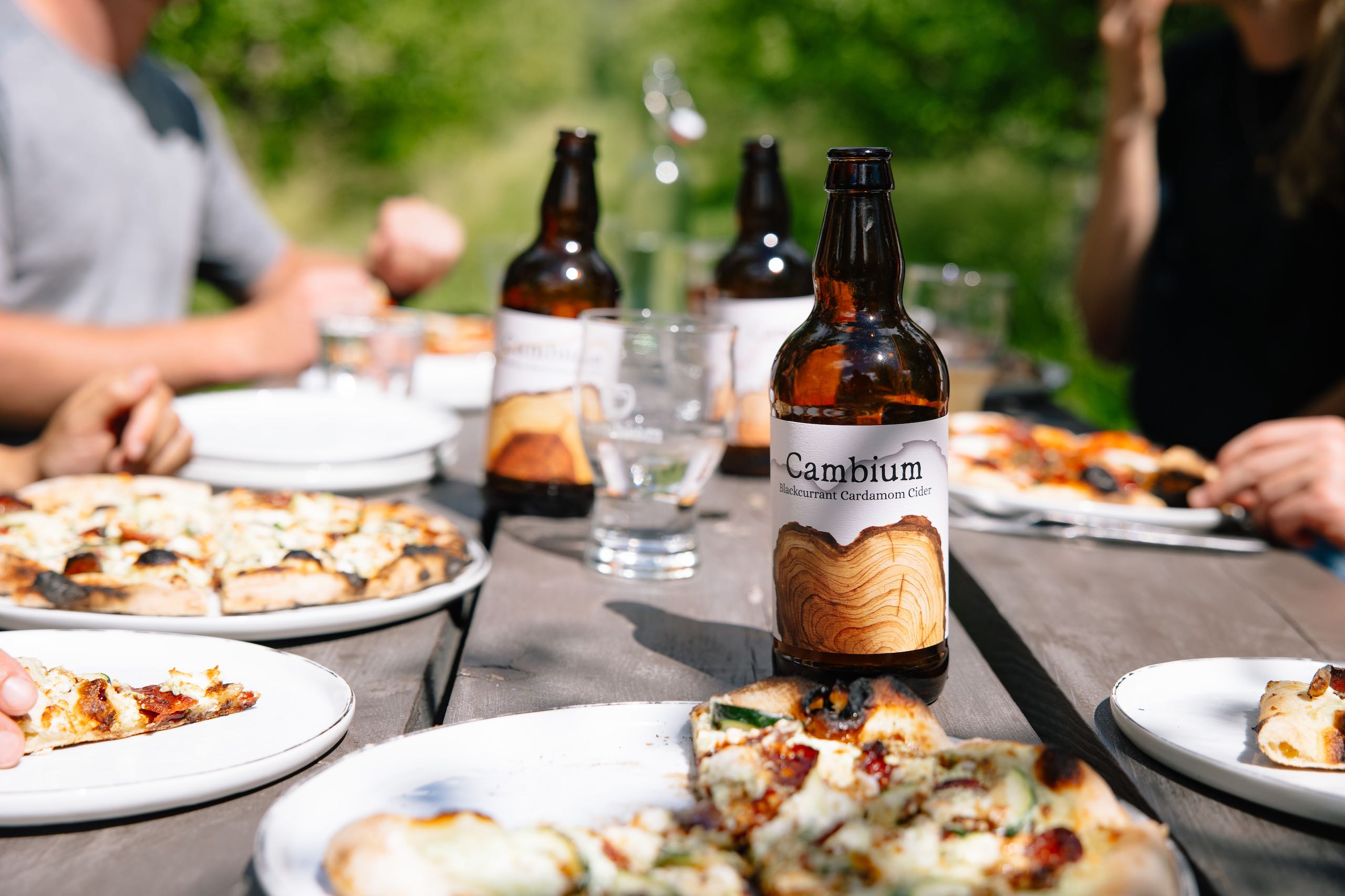 Close up of a Cambium Cider Co. cider bottle on a picnic table with pizza, people, and more cider in the background.