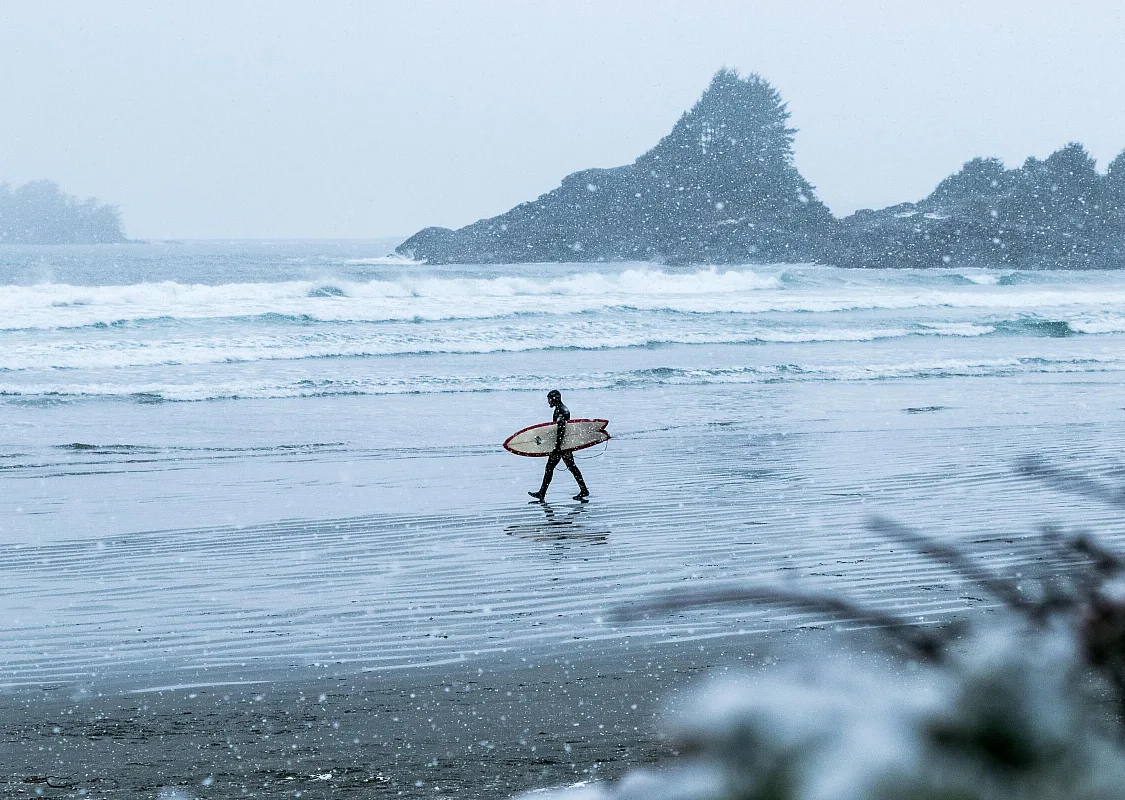 Un surfista camina por la playa hacia el mar, en medio de la nieve, en Cox Bay, Tofino.