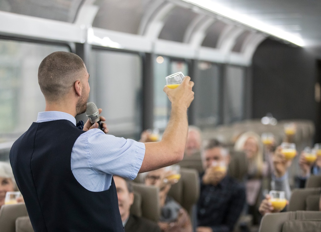 A train host holds a microphone in one had and raises a glass of orange juice in the other, facing a car full of people whose faces are blurry.