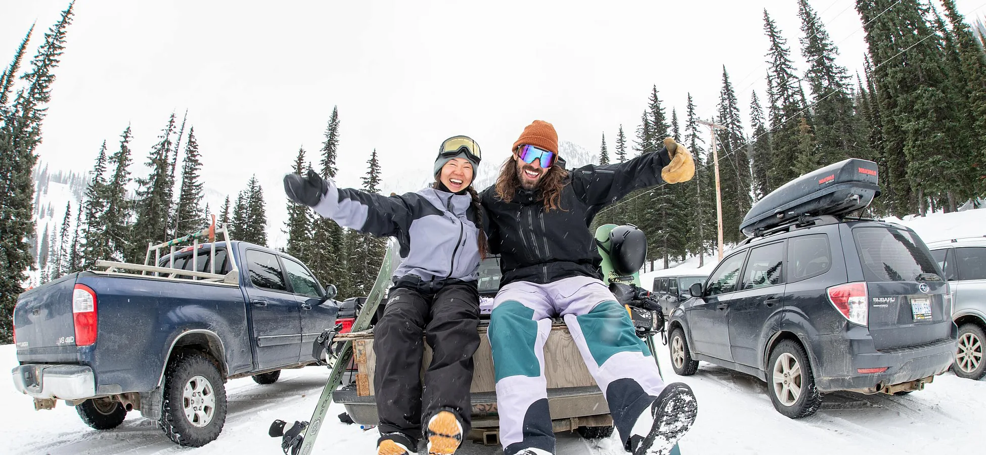 Zwei Snowboarder sitzen auf der Ladefläche eines Pick-ups auf einem verschneiten Parkplatz im Whitewater Mountain Resort und lachen, während ihre Boards zu ihren Füßen liegen.