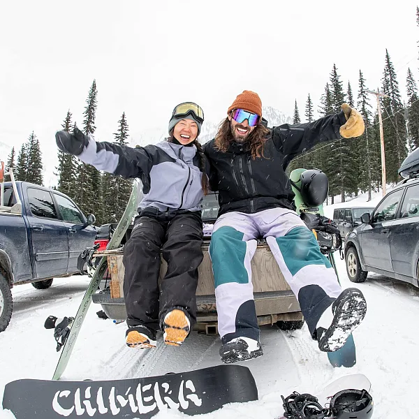 Two snowboarders sitting on the back of a truck in a snowy parking lot at Whitewater Mountain Resort, laughing with their boards at their feet.