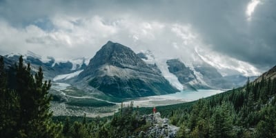A hiker is perched on a trail. In the background is Berg Lake and Mt Robson.