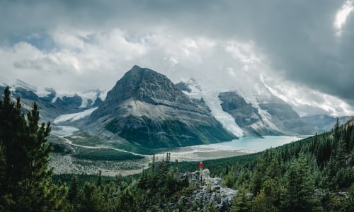 A hiker is perched on a trail. In the background is Berg Lake and Mt Robson.