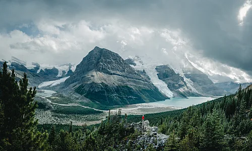 A hiker is perched on a trail. In the background is Berg Lake and Mt Robson.