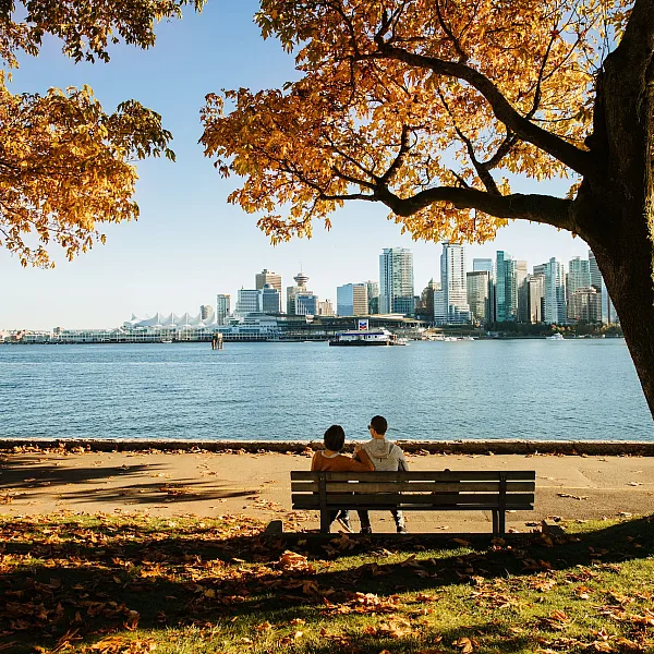 Couple sitting on bench on seawall in Stanley Park looking at downtown skyline in autumn