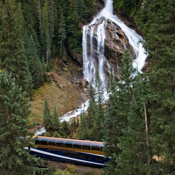 A waterfall down the side of a mountain flows toward a passing train. The waterfall splits near the top with the left side cascading over a ridge and the right curving down the slope.