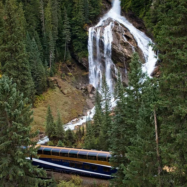 A waterfall down the side of a mountain flows toward a passing train. The waterfall splits near the top with the left side cascading over a ridge and the right curving down the slope.