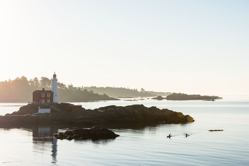 kayaks pass a small island off a rocky shoreline with a white and red lighthouse.