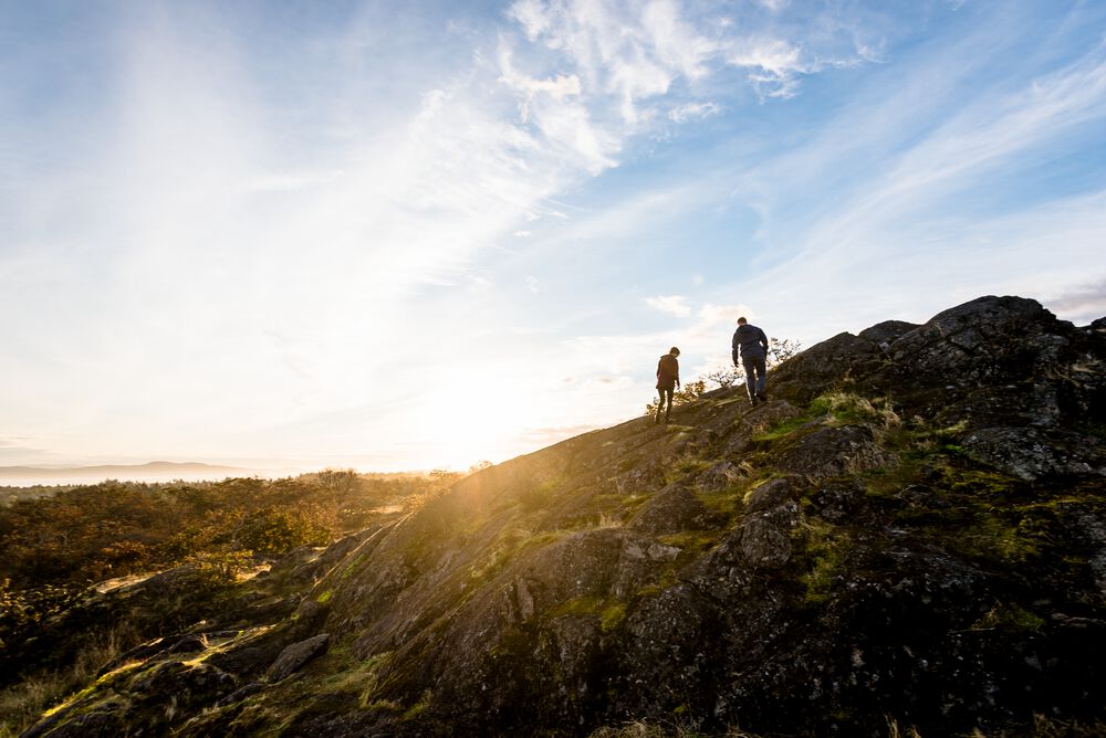 The silhouettes of two people are seen climbing toward the top of a mountain backed by a slightly cloudy blue sky and the sun setting.