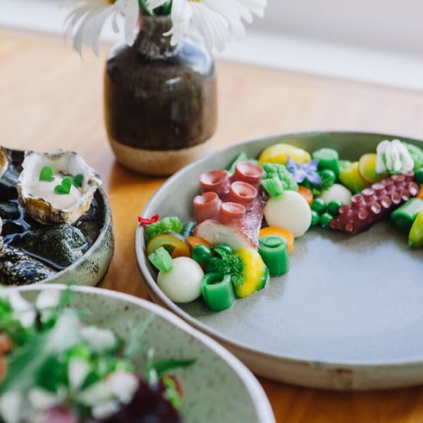 Three plates of food and a small ceramic vase sit on a wooden table. The food includes greens, skinned radishes, and what looks like octopus tentacles.
