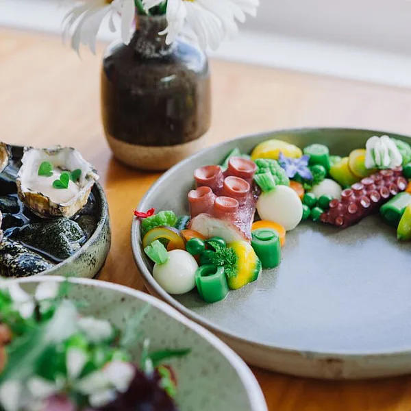 Three plates of food and a small ceramic vase sit on a wooden table. The food includes greens, skinned radishes, and what looks like octopus tentacles.