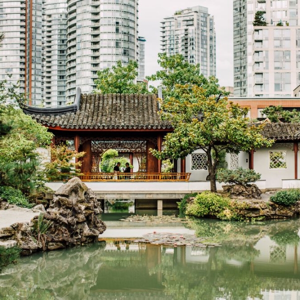 A green pond in the foreground is surrounded by a handful of evergreen trees with a traditional Chinese building behind, and in the distance the towers of downtown Vancouver.