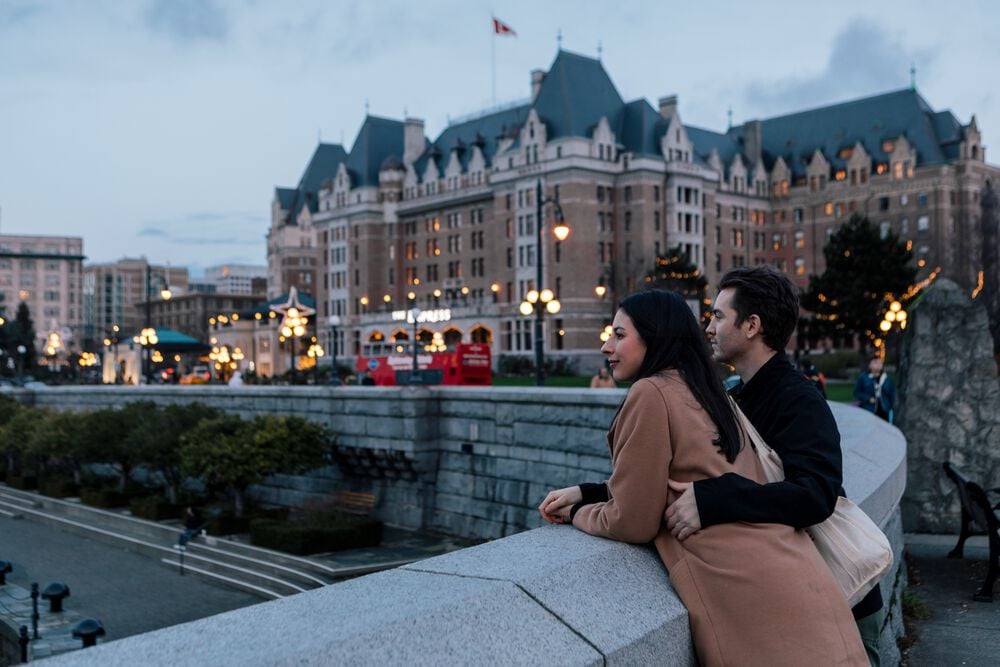 A couple leans against a stone railing overlooking the Victoria Inner Harbour. A stately hotel sits to their right.