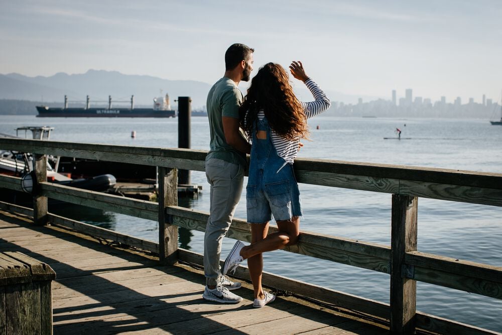 Two people standing on a wooden pier look out over the ocean. A barge is in the water to their left, and in the distance is the downtown Vancouver skyline and the North Shore Mountains,