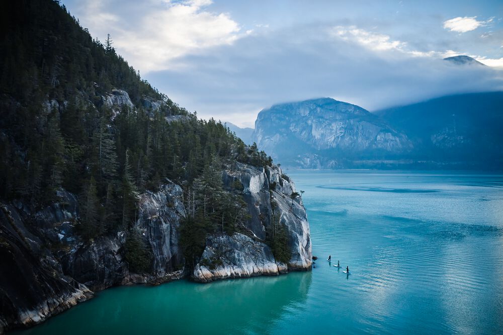 Paddleboarders can be seen on the right of the frame, dwarfed by the huge granite cliffs on Howe Sound.