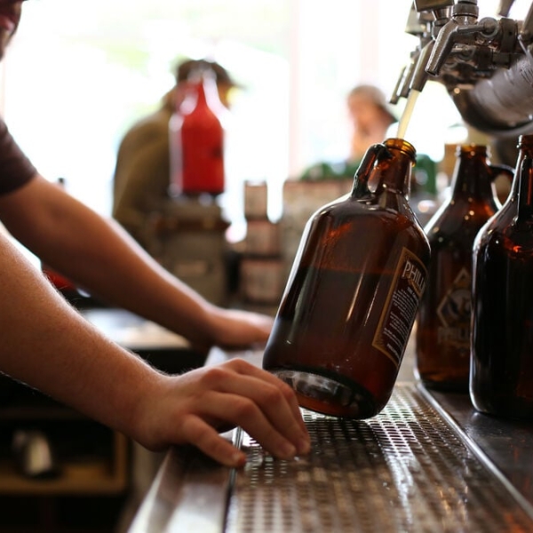 Two arms hold a tilted growler being filled with beer on tap. Empty growlers sit behind.