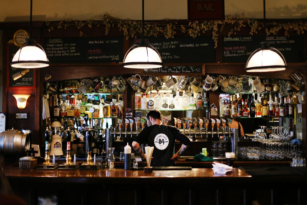 A bartender faces away from the camera toward a series of taps and bottles of alcohol. Mugs hang from above the bar, and there is a chalkboard menu near the cieling.