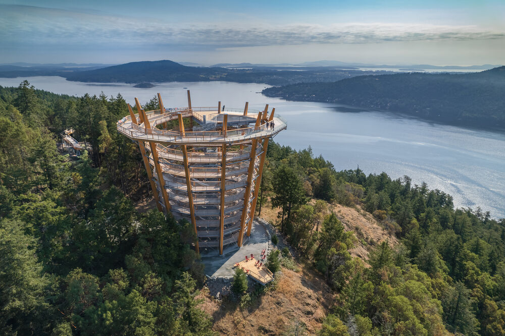 Aerial view of a spiraling ramp that sits in a forested setting overlooking the ocean and islands visible in the background. The ramp gets wider as it gets higher.