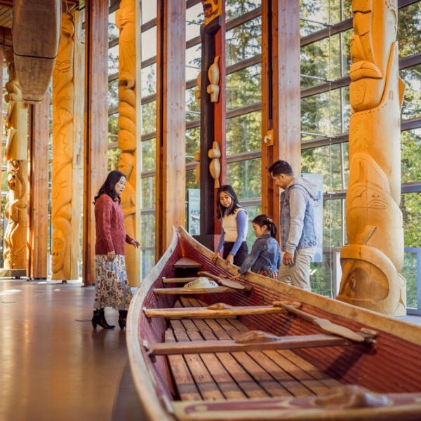 A family of four looks at an Indigenous canoe and carved totem poles at Squamish Lil'wat Cultural Centre.