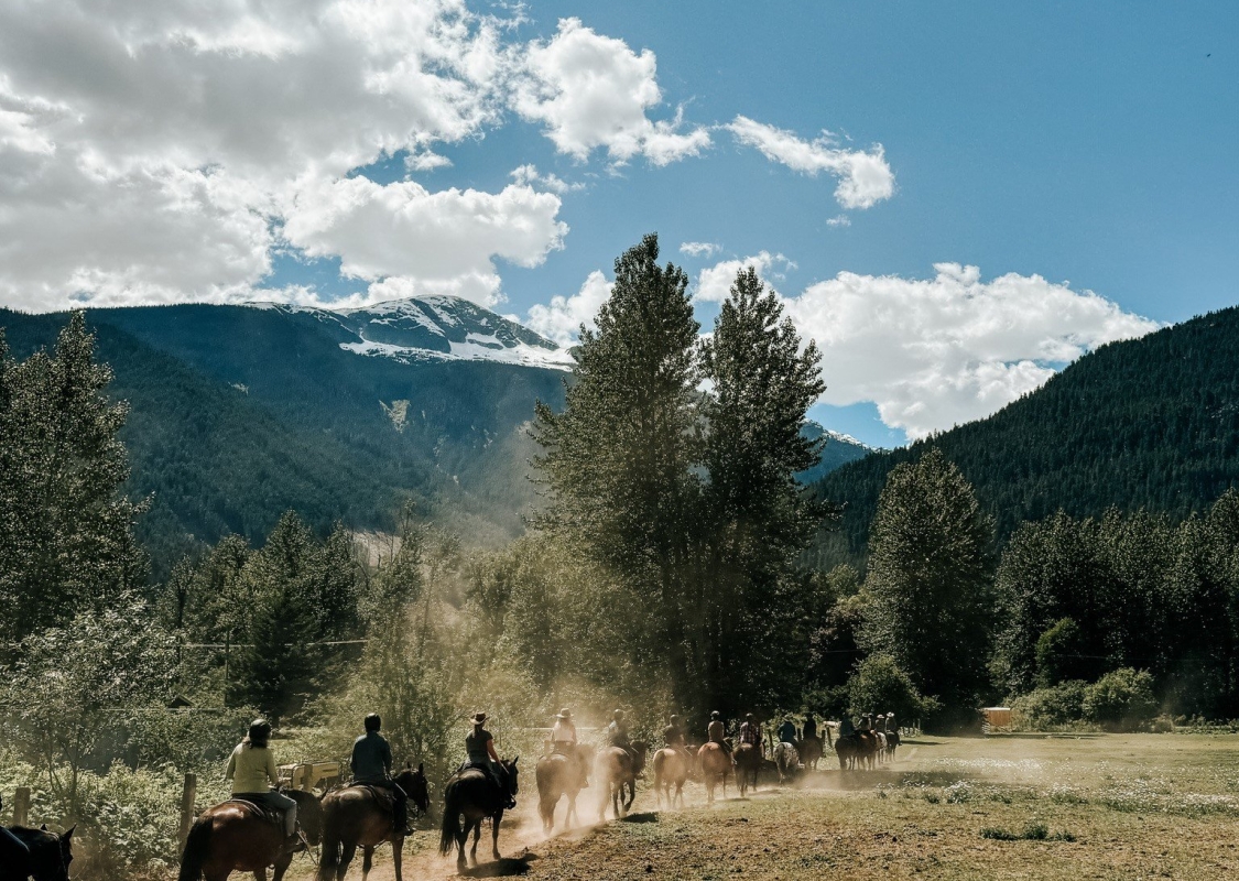 A group of people on horseback walk along a dirt trail with mountains in the background.
