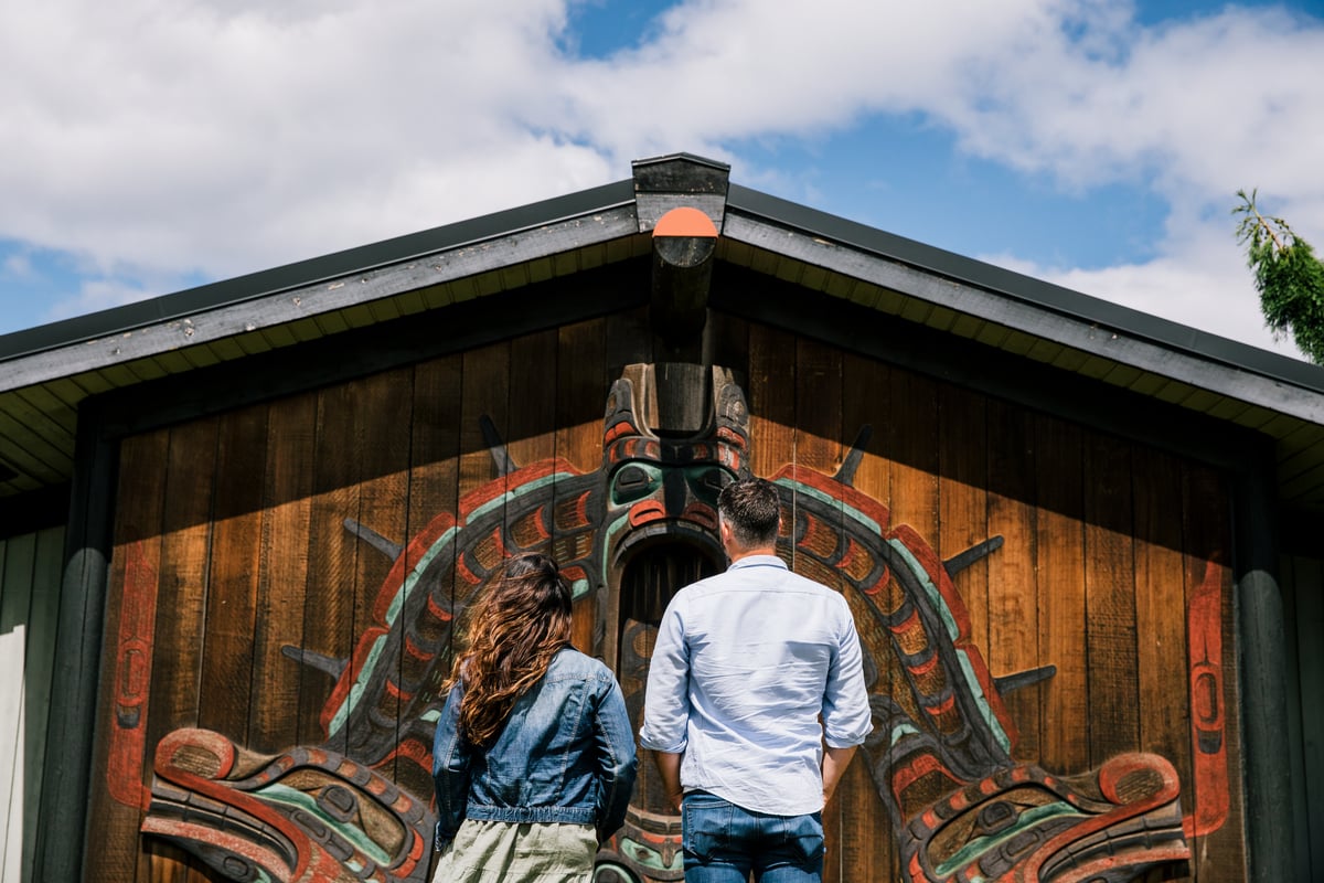 Two people stand in front of a wooden building adorned with a large, intricate Indigenous artwork. The individuals, seen from behind, are looking up at the carving, appreciating its craftsmanship.