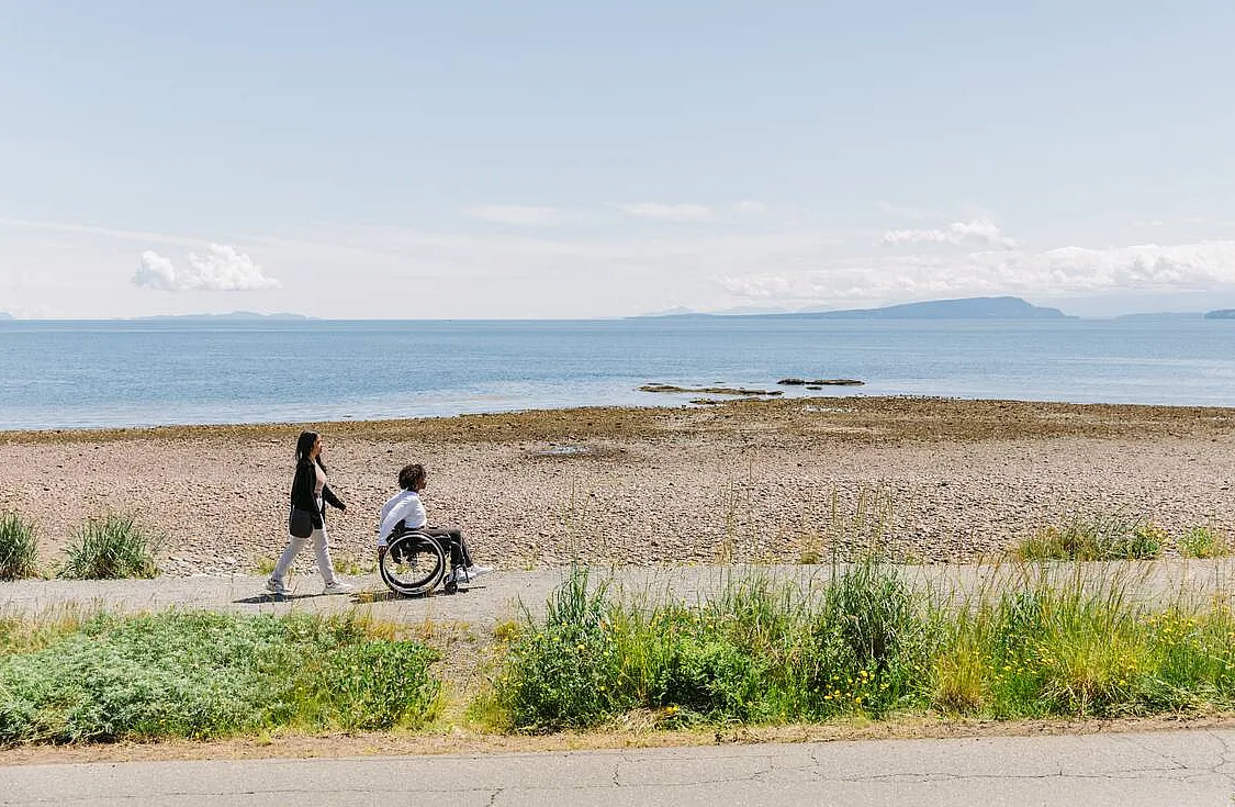 One person is walking and another person uses a wheelchair to go along a seaside accessible trail at Point Holmes near Comox. There is grass in the foreground, and the beach looks out to mountains in the distance.