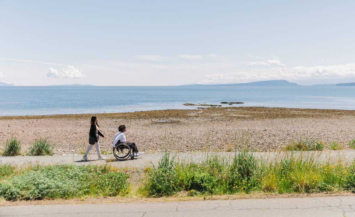 One person is walking and another person uses a wheelchair to go along a seaside accessible trail at Point Holmes near Comox. There is grass in the foreground, and the beach looks out to mountains in the distance.