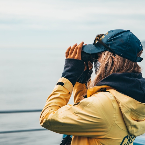 A guide wearing a yellow rain jacket and a blue cap on a Prince of Whales tour scans the water through a pair of binoculars