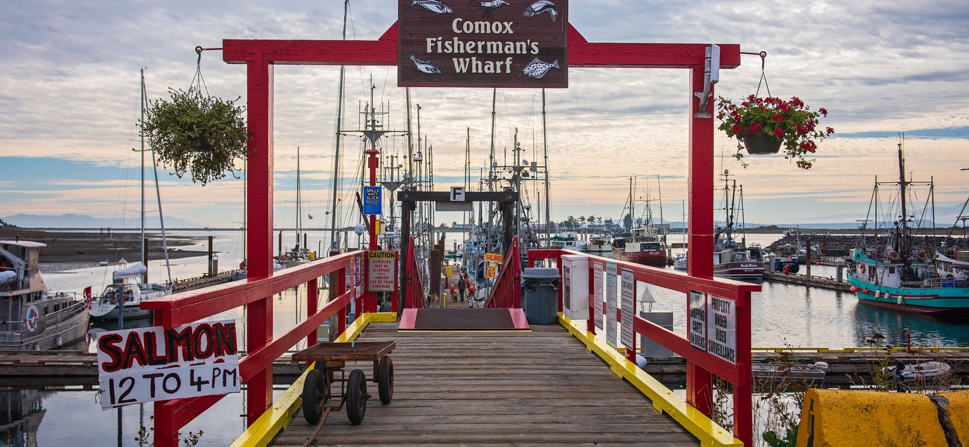 Brightly-coloured red and yellow archway and railing leading out to a dock lined with fishing vessels. A sign reads, 