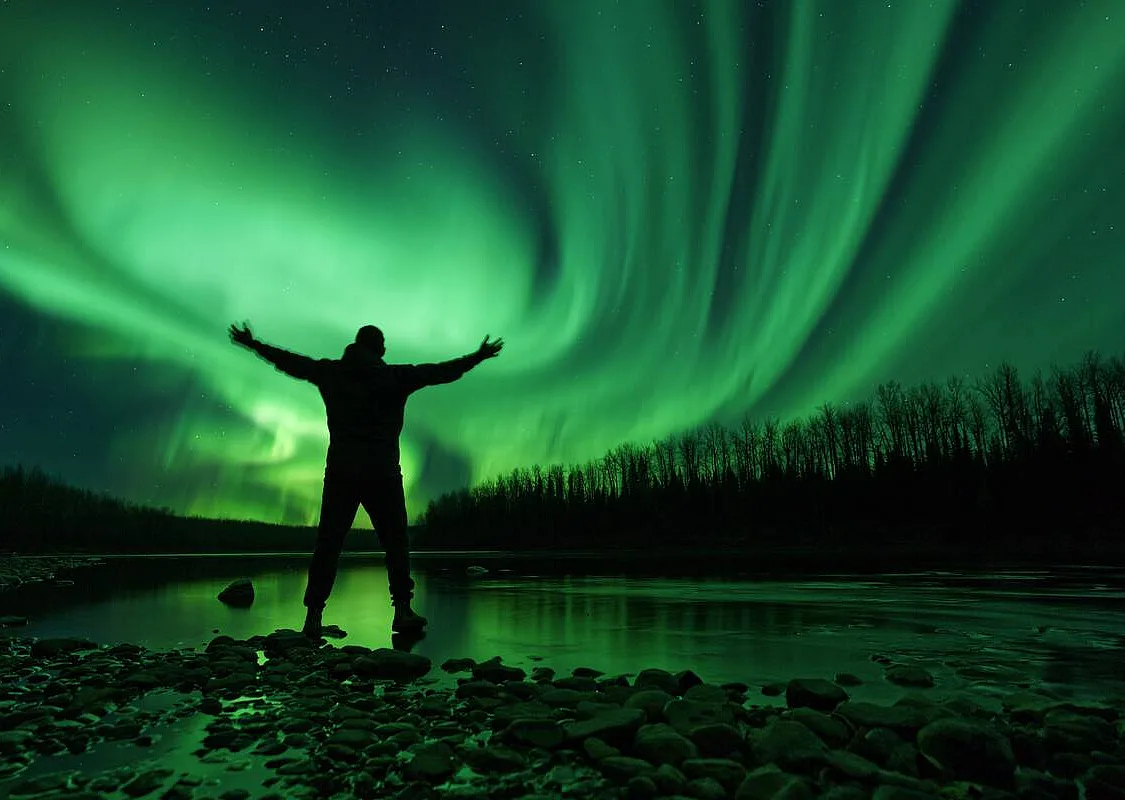 A silhouette of a person stands with their arms outstreached to the sky as neon green Northern Lights dance across the landscape.