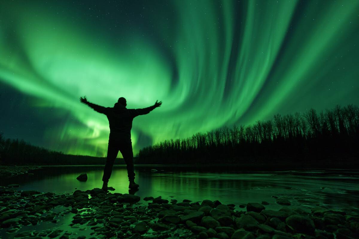 A silhouette of a person stands with their arms outstreached to the sky as neon green Northern Lights dance across the landscape.