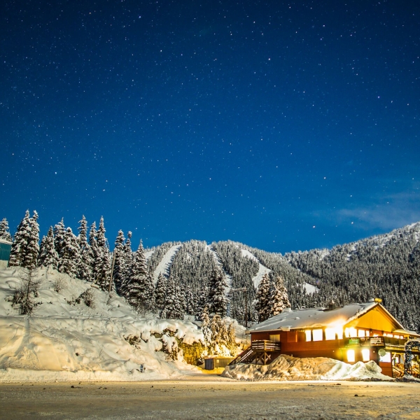 A wooden lodge sits in a gorgeous snowy setting with trees and mountains behind. It's dusk, and the lights are on inside the lodge.