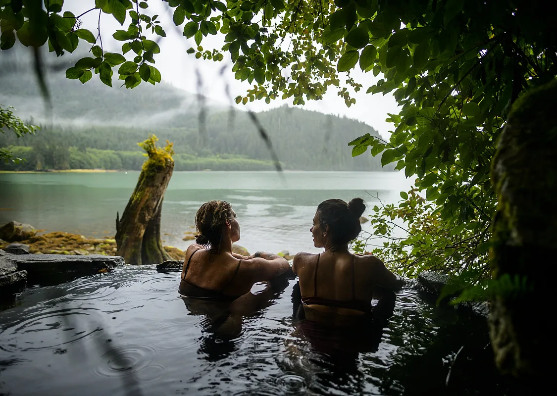 Two people soak in a natural hot springs pool with misty rainforest, mountains and waterways in the distance.