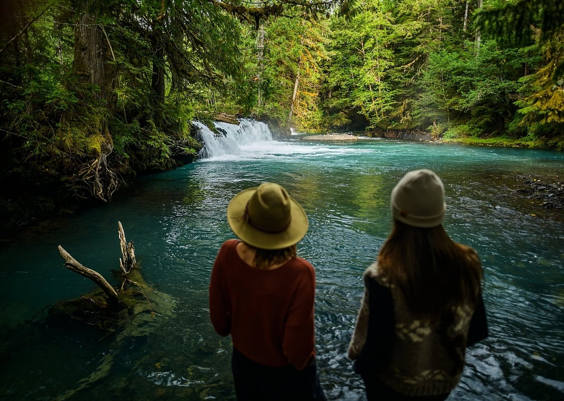 Zwei Personen blicken auf einen türkisfarbenen Wasserfall, umgeben von dichtem grünem Wald.