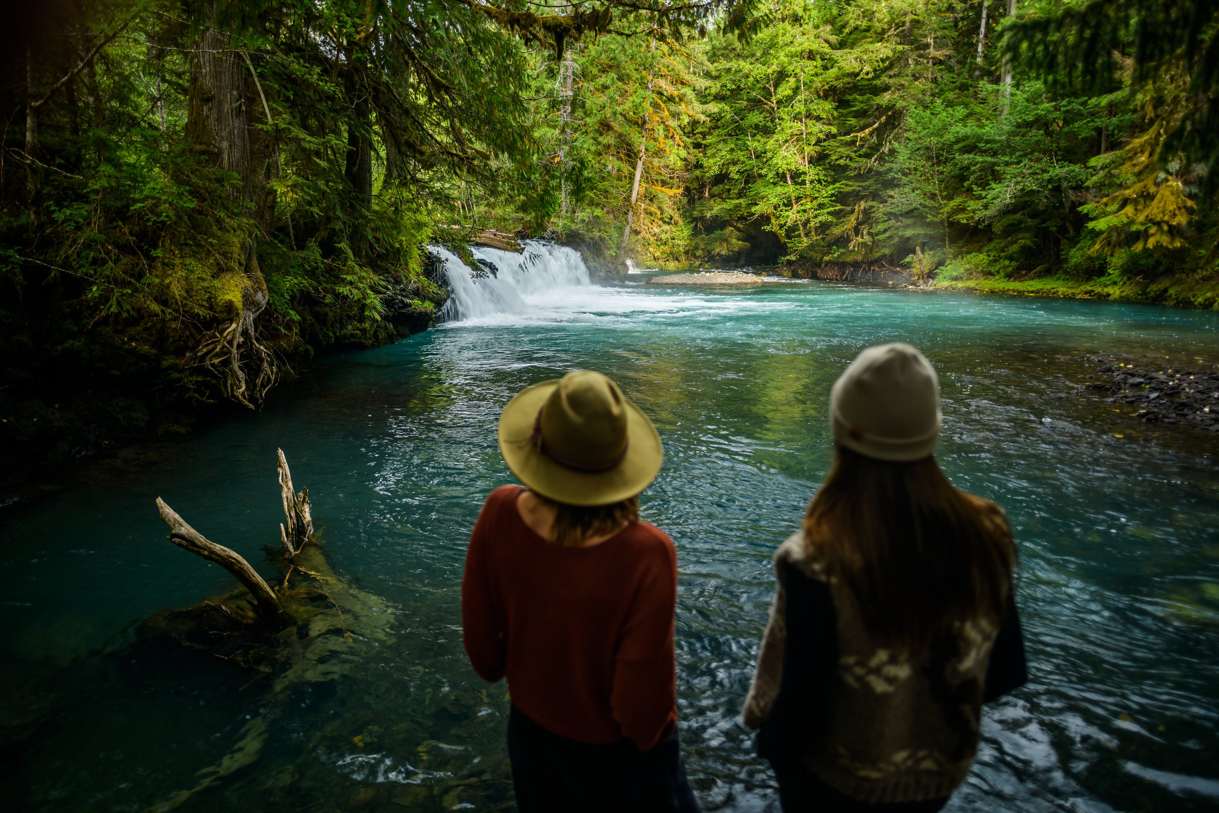 Two people look out over a turquoise waterfall surrounded by dense green forest.