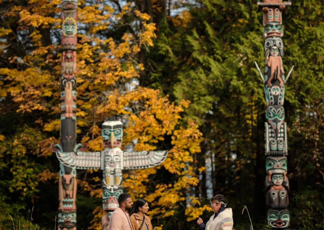 Two people listen to a guide dressed in Indigenous attire; the group is standing in front of three totems set in a forest.