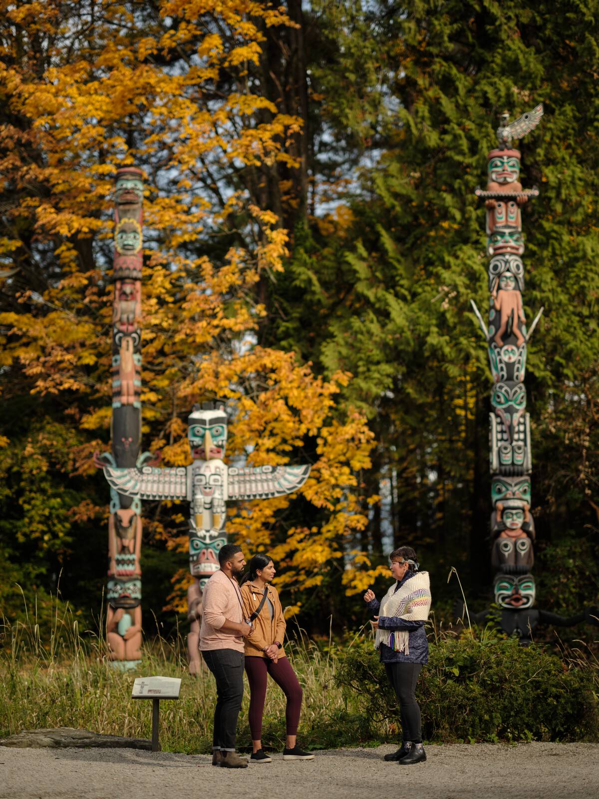 Two people listen to a guide dressed in Indigenous attire; the group is standing in front of three totems set in a forest.