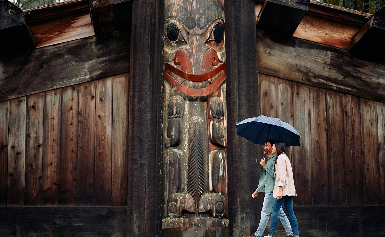 Zwei Leute gehen unter einem blauen Regenschirm, mit einem verwitterten indigenen Pfahl und einem Gebäude hinter ihnen.