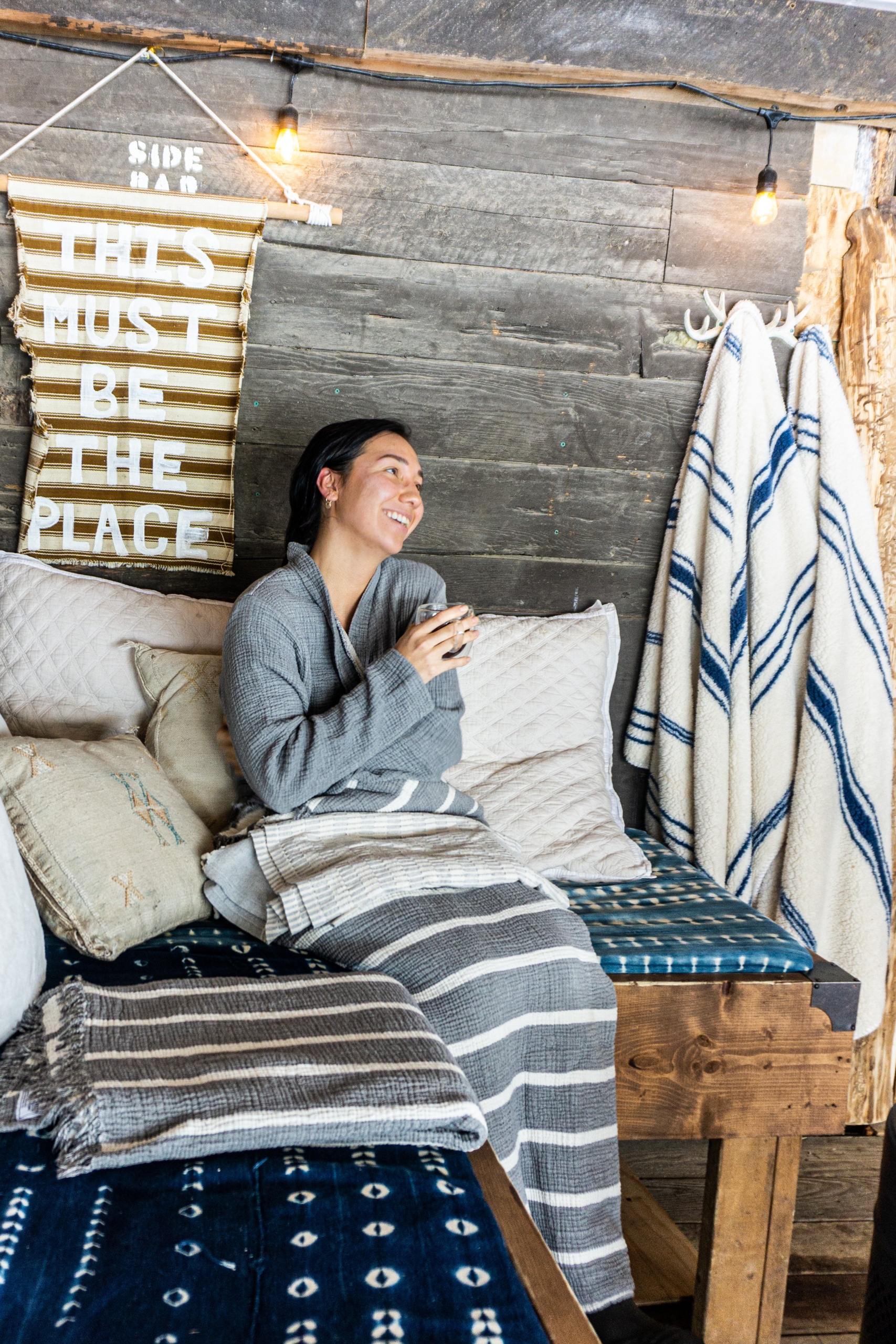 Person wrapped in a grey and white striped blanket, backed by pillows, holds a beverage while sitting on an indoor wooden bench