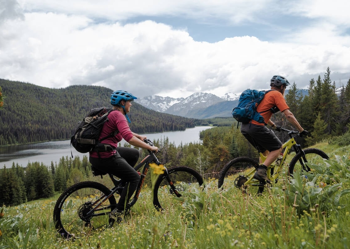 Two mountain bikers ride up a trail through a grassy meadow overlooking a lake surrounded by forest, with mountains in the background.