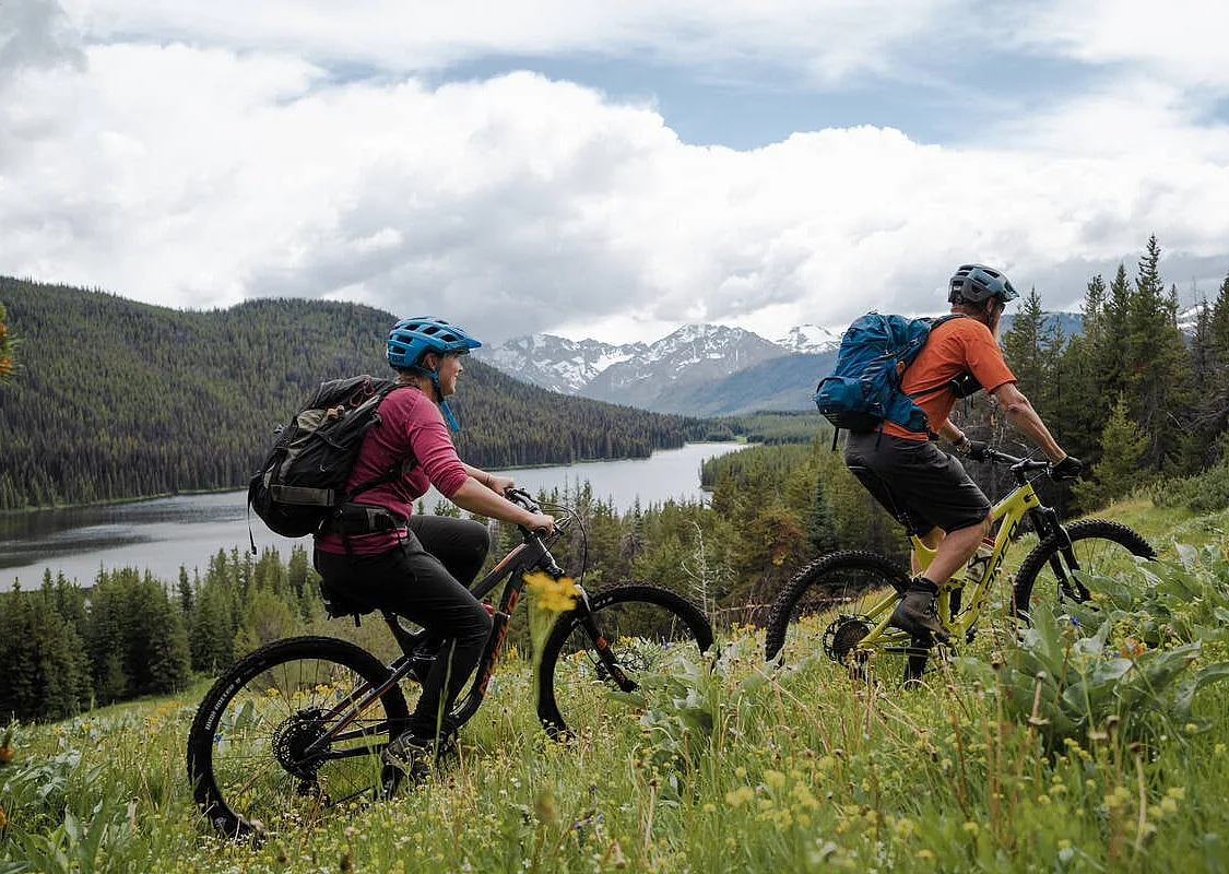 Two mountain bikers ride up a trail through a grassy meadow overlooking a lake surrounded by forest, with mountains in the background.
