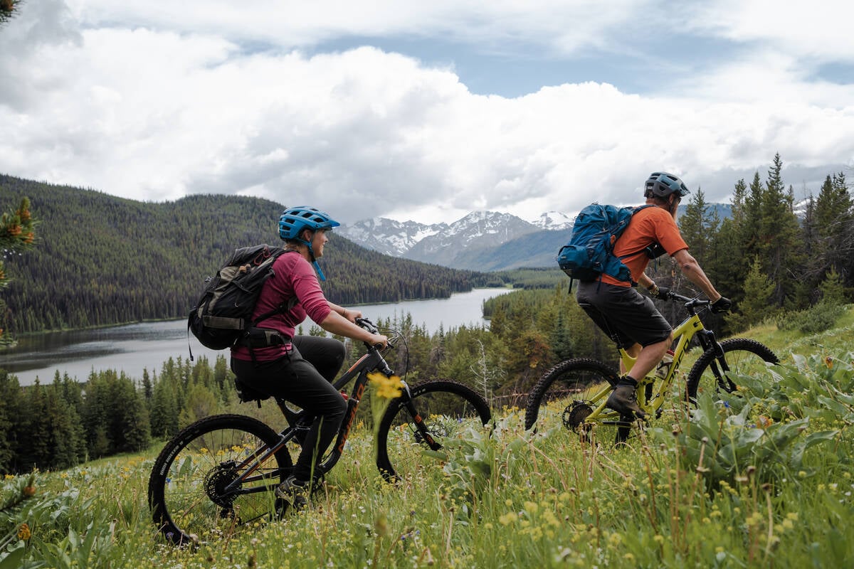 Two mountain bikers ride up a trail through a grassy meadow overlooking a lake surrounded by forest, with mountains in the background.