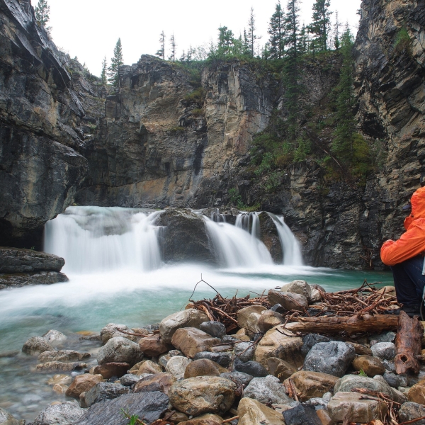 A person sits on a rocky shore looking out at a small waterfall in a canyon.