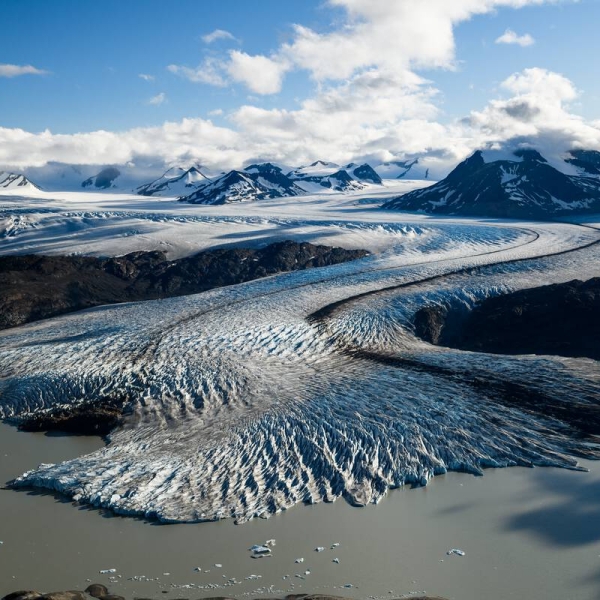 A snaking glacier with white and blue ice snakes across an icefield, dotted by mountains and a glacial lake.