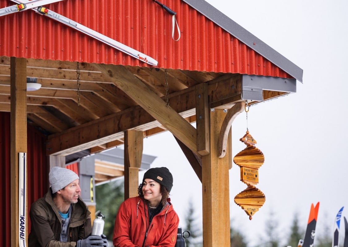 Two people stand on the deck of a wooden building with red siding. The railing on the deck is made from colourful skis, and there are white, criss-crossed skis above the deck.