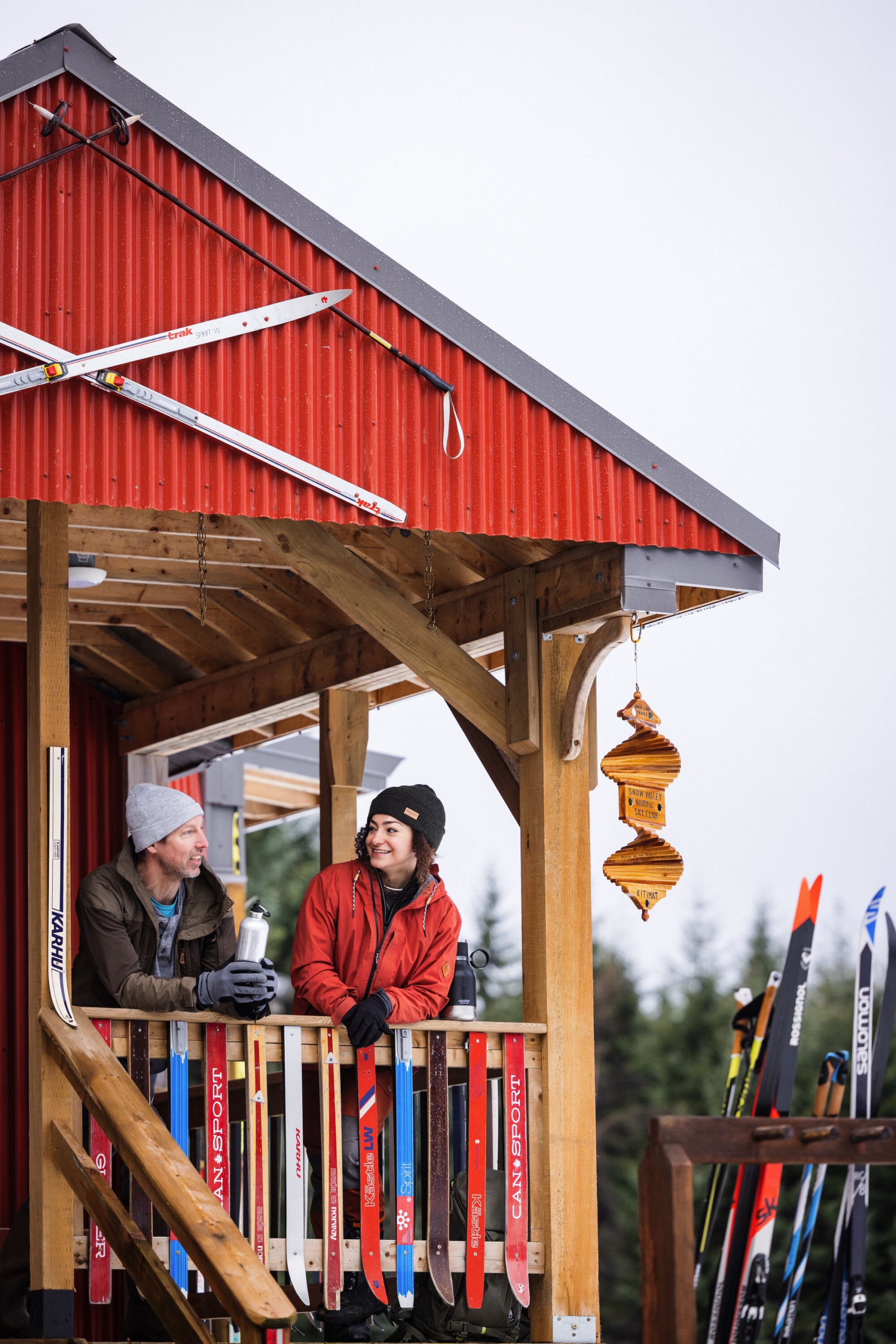 Two people stand on the deck of a wooden building with red siding. The railing on the deck is made from colourful skis, and there are white, criss-crossed skis above the deck.