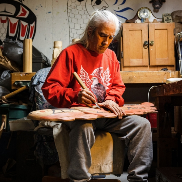 An Indigenous artist in a red sweatshirt sits in a chair in what looks to be his workshop working on a long, flat carving.