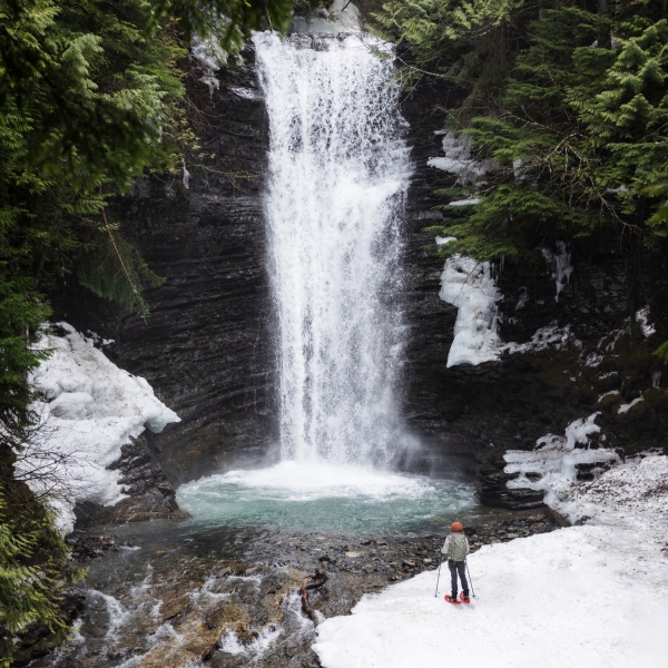 A person stands on the snowy banks of a body of water as a short waterfall plummets into a pool.