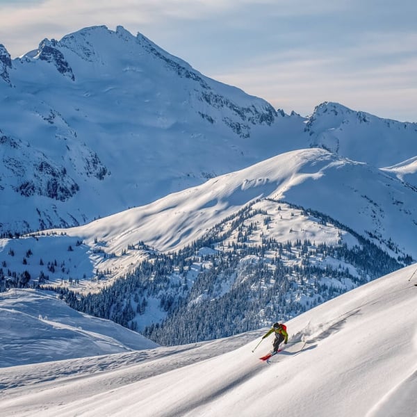 two skiers descend through the powder on a steep slope, several mountain peaks behind them